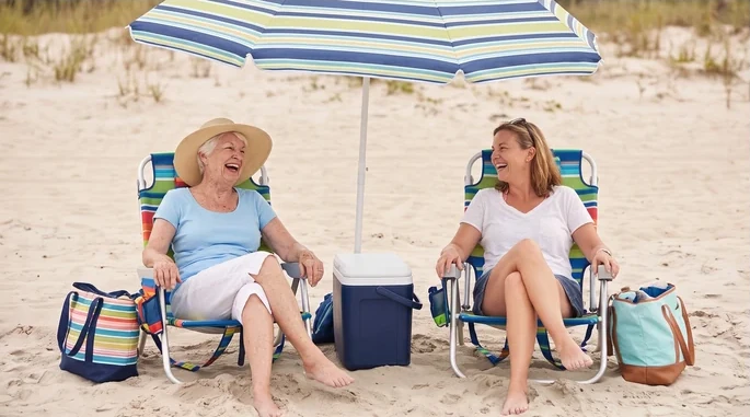 Ruth, a white-haired woman in her seventies in a sun hat, laughing with her grown daughter in beach chairs under a striped umbrella
